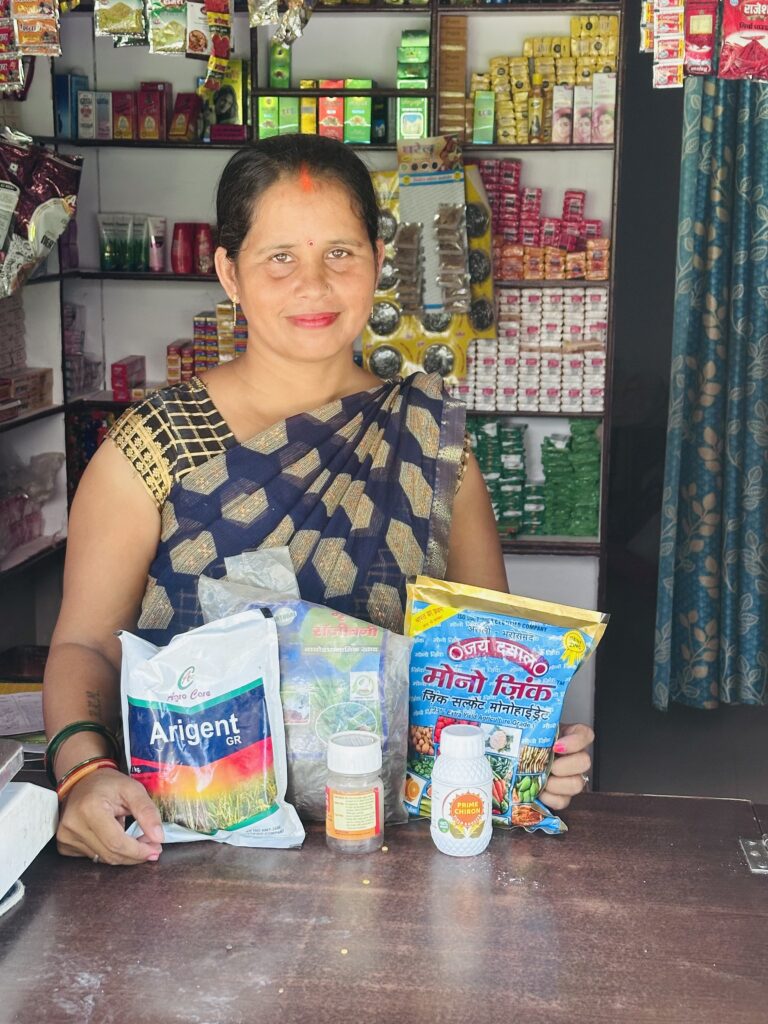 Sanju Maurya's Agri-Entrepreneur role with Udhyami Mahila FPC in her shop standing at counter showing some of the products she sells